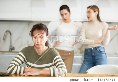 Upset middle-aged woman sitting at the kitchen table with her back to women quarreling to her 138578254