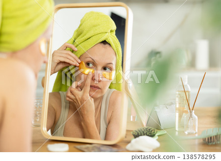 Woman in towel turban applying golden collagen eye patches Woman in towel turban applying golden collagen eye patches 138578295