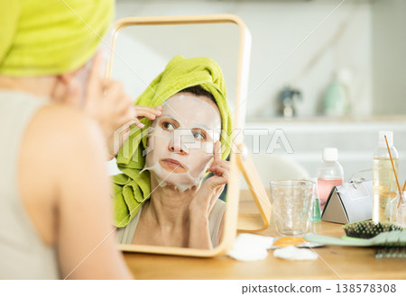 Woman applying moisturizing sheet face mask in front of mirror Woman applying moisturizing sheet face mask in front of mirror 138578308