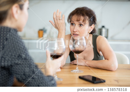 Sad middle-aged woman sitting at table with a glass of wine and talking to a friend Sad middle-aged woman sitting at table with a glass of wine and talking to a friend 138578318