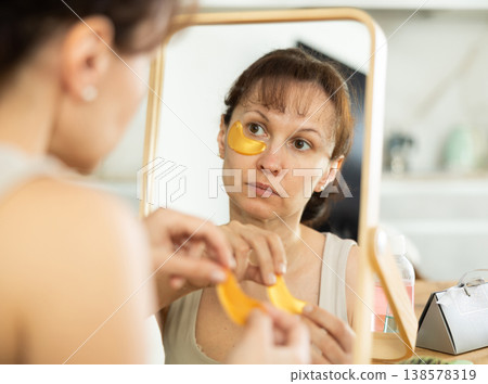 Woman using gold collagen eye patches in front of mirror Woman using gold collagen eye patches in front of mirror 138578319