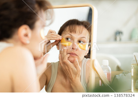 Middle-aged woman applying eye patch mask on her face sitting in front of the mirror Middle-aged woman applying eye patch mask on her face sitting in front of the mirror 138578324