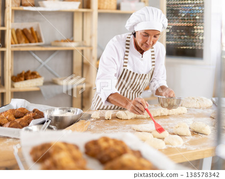 Aged woman baker buttering pieces of dough in bakery Aged woman baker buttering pieces of dough in bakery 138578342