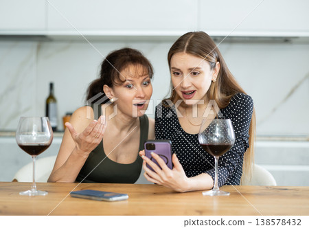 Two happy middle-aged women looking at the phone display while sitting at the kitchen-table Two happy middle-aged women looking at the phone display while sitting at the kitchen-table 138578432