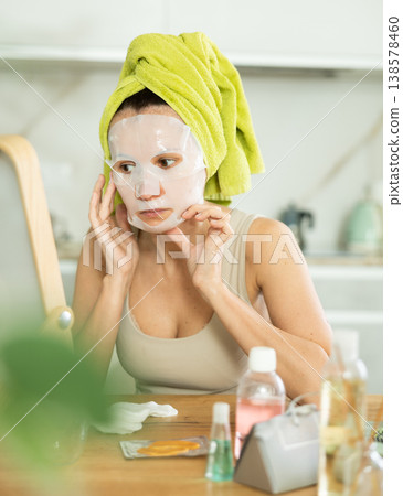 Middle-aged woman applying tissue mask on her facial skin sitting in front of the mirror 138578460