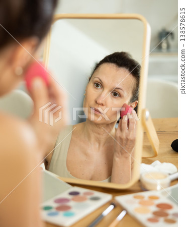 Middle-aged woman applying make-up foundation on her face by means of sponge Middle-aged woman applying make-up foundation on her face by means of sponge 138578615