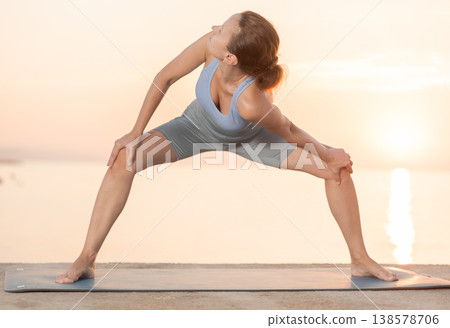 Young positive coach female in activewear doing stretching exercises during training yoga on seashore at sunrise 138578706
