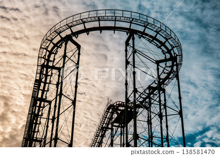 Dark silhouette of a roller coaster track against a bright, textured cloud background at dusk Dark silhouette of a roller coaster track against a bright, textured cloud background at dusk 138581470
