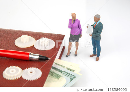 Miniature people talk over gears and money in a business setting with a pen and notebook Miniature people talk over gears and money in a business setting with a pen and notebook 138584093