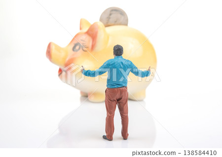 Miniature person stands near a large piggy bank with arms raised while facing the bank  138584410