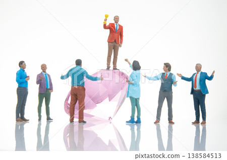 Miniature people celebrate on a large gem while one holds a trophy under bright light Miniature people celebrate on a large gem while one holds a trophy under bright light 138584513