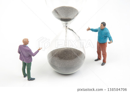 Miniature people talk by an hourglass filled with dark sand in a simple setting Miniature people talk by an hourglass filled with dark sand in a simple setting 138584706