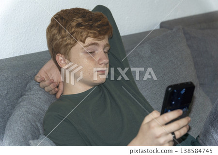 Teenage boy with red hair sitting on a sofa and using his smartphone. Teenage boy with red hair sitting on a sofa and using his smartphone. 138584745