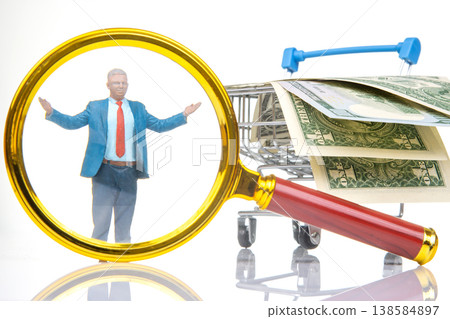 Miniature people in a shopping cart surrounded by money under a magnifying glass in a bright setting Miniature people in a shopping cart surrounded by money under a magnifying glass in a bright setting 138584897