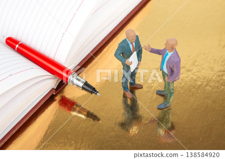 Miniature figurines have a discussion next to a large pen and an open planner on a shiny surface Miniature figurines have a discussion next to a large pen and an open planner on a shiny surface 138584920