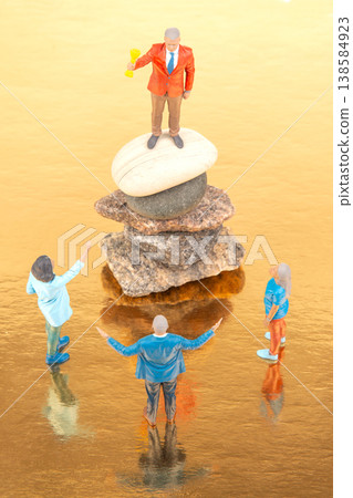 Miniature people stand on stones while one figure holds a trophy above others in a water setting Miniature people stand on stones while one figure holds a trophy above others in a water setting 138584923
