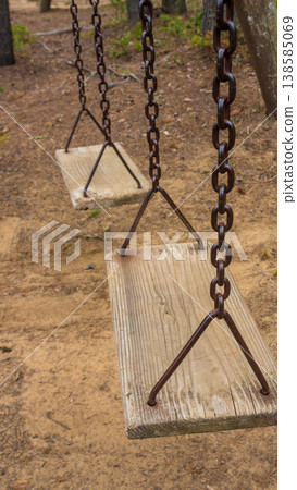 Wooden swings hanging from chains in a forest playground area with sandy ground and trees in the background Wooden swings hanging from chains in a forest playground area with sandy ground and trees in the background 138585069