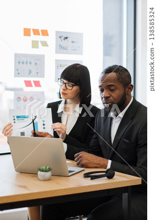 A woman in glasses explains a graph to an African American man during a business meeting, with a laptop and charts on the table 138585324