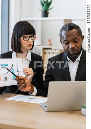 A woman points at a financial chart while discussing with an African American man during a business meeting A woman points at a financial chart while discussing with an African American man during a business meeting 138585332