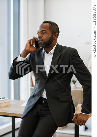 A professional African American man in a suit talks on his mobile phone while leaning on a desk in a modern office setting A professional African American man in a suit talks on his mobile phone while leaning on a desk in a modern office setting 138585378