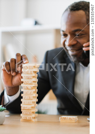 An African American man in a suit plays a game of wooden blocks while talking on the phone, demonstrating focus and multitasking An African American man in a suit plays a game of wooden blocks while talking on the phone, demonstrating focus and multitasking 138585467