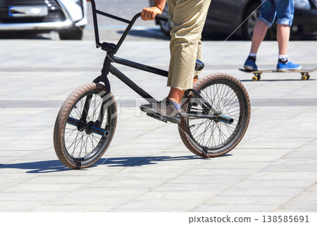 Biker rides a black BMX bike on pavement while a person skateboards in the background during Biker rides a black BMX bike on pavement while a person skateboards in the background during 138585691
