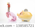 Miniature people stand on a crystal holding a trophy, while a piggy bank waits with a coin resting  138585721