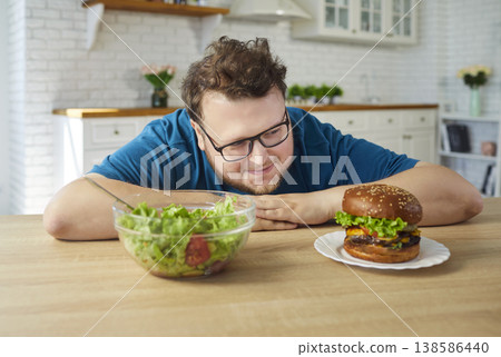 Obese man choosing salad or juicy burger at home kitchen, diet temptation Obese man choosing salad or juicy burger at home kitchen, diet temptation 138586440