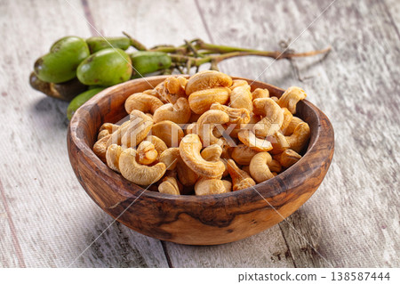 Roasted cashew nuts in a brown bowl with a cluster of raw green cashew fruit on a rustic wooden table. 138587444
