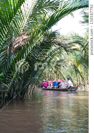 Tourists in conical hats enjoy a boat ride through lush green palm trees along a river in the Mekong Delta 138587792