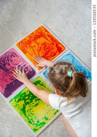Little girl pressing colorful sensory gel mats with both hands during tactile play activity at home. Sensory integration, autism support and occupational therapy development concept. Little girl pressing colorful sensory gel mats with both hands during tactile play activity at home. Sensory integration, autism support and occupational therapy development concept. 138588535