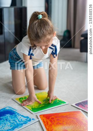 Little girl kneeling on floor pressing colorful sensory gel mats with hands during play activity. Sensory stimulation, autism support and occupational therapy development concept. Little girl kneeling on floor pressing colorful sensory gel mats with hands during play activity. Sensory stimulation, autism support and occupational therapy development concept. 138588536