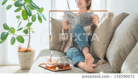 一位穿著時尚的亞洲女性坐在客廳的沙發上,在下午茶時間閱讀雜誌。 一位穿著時尚的亞洲女性坐在客廳的沙發上,在下午茶時間閱讀雜誌。 138590072