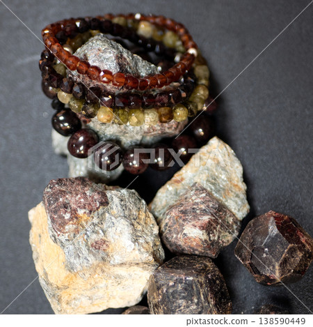 Handcrafted almandine garnet bracelets and raw mineral specimens resting on dark surface. Polished gemstone beads and natural rough stones Handcrafted almandine garnet bracelets and raw mineral specimens resting on dark surface. Polished gemstone beads and natural rough stones 138590449