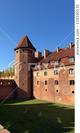 Malbork Castle in Poland with round brick tower and defensive walls surrounding green courtyard under clear blue sky. Architecture, history and travel concept 138590530