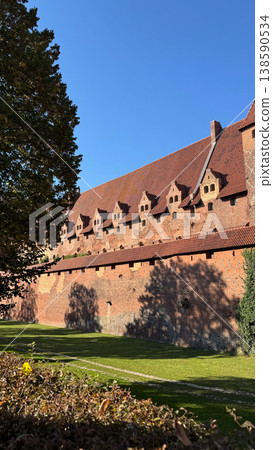 Malbork Castle in Poland with extended red brick wall and repeated roof dormers under clear blue sky with tree shadows on facade. Architecture, history and travel concept Malbork Castle in Poland with extended red brick wall and repeated roof dormers under clear blue sky with tree shadows on facade. Architecture, history and travel concept 138590534
