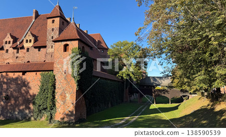 Malbork Castle in Poland with red brick walls tower and wooden bridge over grassy moat with trees under blue sky. Architecture, history and travel concept Malbork Castle in Poland with red brick walls tower and wooden bridge over grassy moat with trees under blue sky. Architecture, history and travel concept 138590539