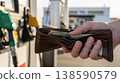 Close-up of a hand holding a leather wallet with cash at a gas station pump, highlighting fuel expenses, everyday spending, and the rising cost of travel and transportation 138590579