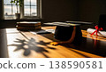 A black graduation cap rests next to a diploma tied with a red ribbon on a wooden table shaded by a window. The academic background symbolizes success, goal achievement, and graduation. 138590581
