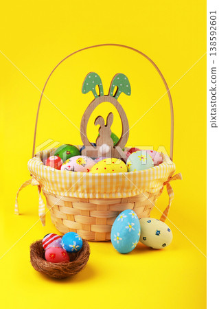 Colorful hand-painted Easter eggs in a wicker basket with a wooden bunny ornament and a small nest against a vibrant yellow background. Cheerful spring holiday decoration. High quality photo 138592601