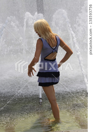 Young blonde hair woman, stands barefoot in a shallow fountain,,surrounded by splashing water jets. 138593756