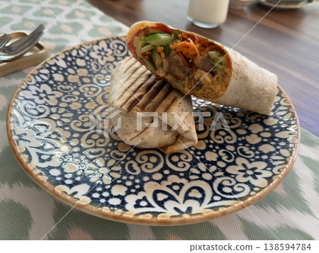 Grilled wrap with vegetables and meat on decorative plate, placed on a wooden table. Grilled wrap with vegetables and meat on decorative plate, placed on a wooden table. 138594784