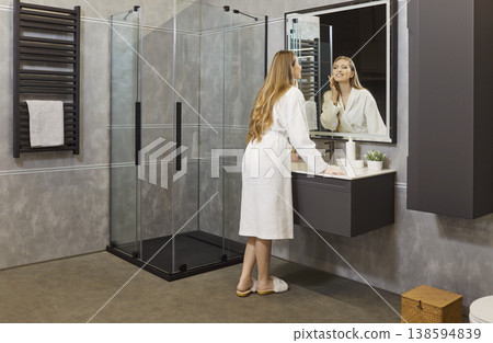 Cheerful young woman standing in modern bathroom and looking at mirror enjoying morning routine. Cheerful young woman standing in modern bathroom and looking at mirror enjoying morning routine. 138594839