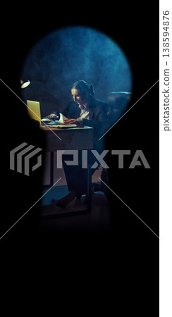 Businesswoman working on laptop at desk in dark room seen through keyhole. 138594876