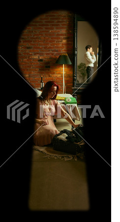 Keyhole view of woman in dress packing suitcase on floor in bedroom with man reflection in mirror. Keyhole view of woman in dress packing suitcase on floor in bedroom with man reflection in mirror. 138594890