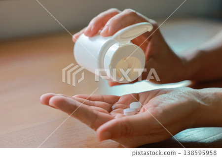 Close up of person hands pouring white pills from a white plastic bottle into their palm. Taking daily medicine, vitamin, painkiller or dietary supplement for healthcare medical treatment concept. Close up of person hands pouring white pills from a white plastic bottle into their palm. Taking daily medicine, vitamin, painkiller or dietary supplement for healthcare medical treatment concept. 138595958