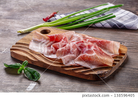 Thinly sliced cured meat, possibly prosciutto, on a wooden cutting board with green onions and basil. Thinly sliced cured meat, possibly prosciutto, on a wooden cutting board with green onions and basil. 138596405