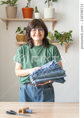 Smiling adult woman standing, holding a stack of old denim jeans for cutting and sewing, promoting sustainable clothing reuse. Environmental awareness Smiling adult woman standing, holding a stack of old denim jeans for cutting and sewing, promoting sustainable clothing reuse. Environmental awareness 138596513