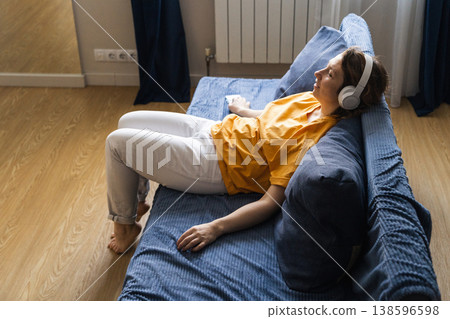 Woman lying on couch wearing white wireless headphones, enjoying audio from smartphone, surfing the internet in a cozy living room 138596598