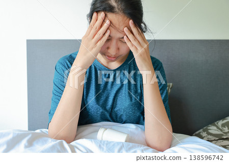 Sick Asian woman sitting on bed holding her head in pain with a white pill bottle on blanket. Concept of severe headache, migraine, stress, insomnia, depression, healthcare and medical treatment. Sick Asian woman sitting on bed holding her head in pain with a white pill bottle on blanket. Concept of severe headache, migraine, stress, insomnia, depression, healthcare and medical treatment. 138596742